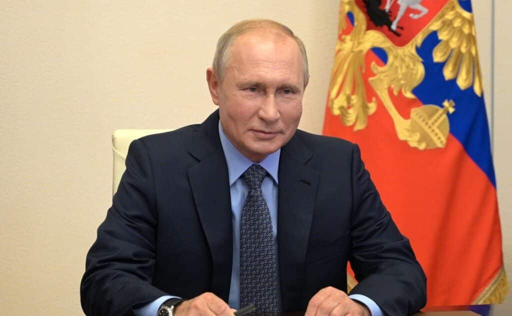 Vladimir Putin, homem de pele clara, poucos cabelos loiros, veste terno e gravata. Atrás dele aparece parte de uma bandeira.