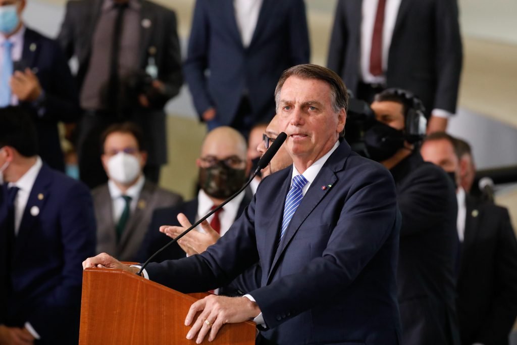 Jair Bolsonaro, de terno escuro e gravata azul, fala ao microfone enquanto outros homens de terno e gravata ao fundo acompanham.