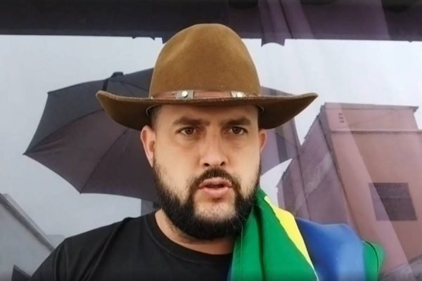 Zé Trovão, homem branco, com bigode e barba, usa chapéu marrom, a bandeira do Brasil sobre o ombro esquerdo e veste camiseta preta.