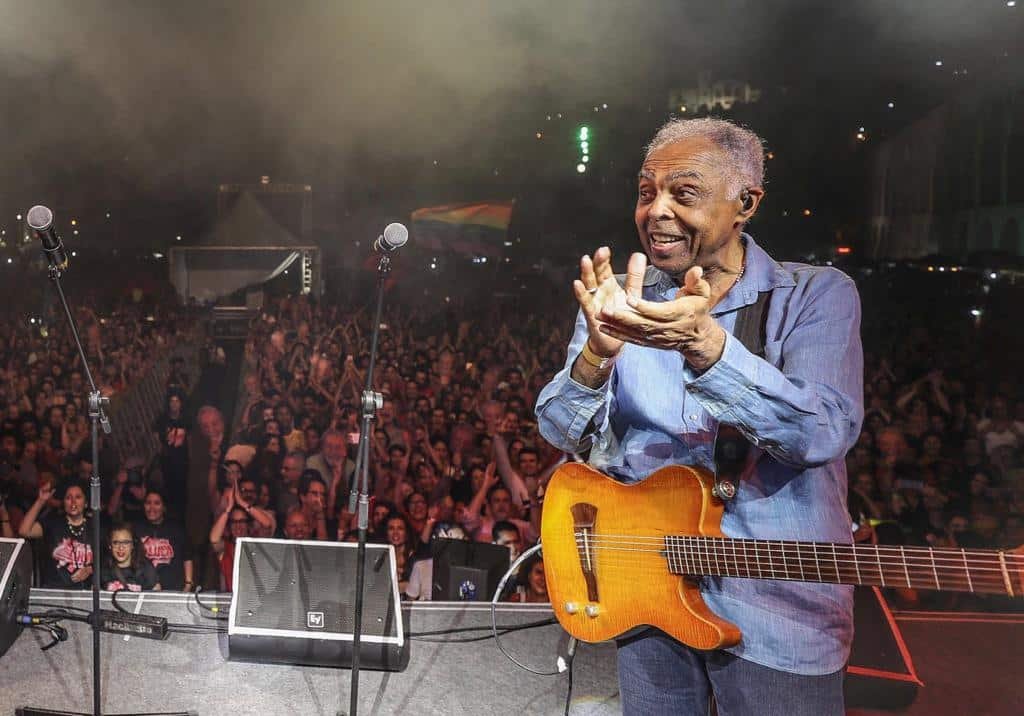 Gilberto Gil no palco, diante do público, batendo palma com o violão preso ao corpo.