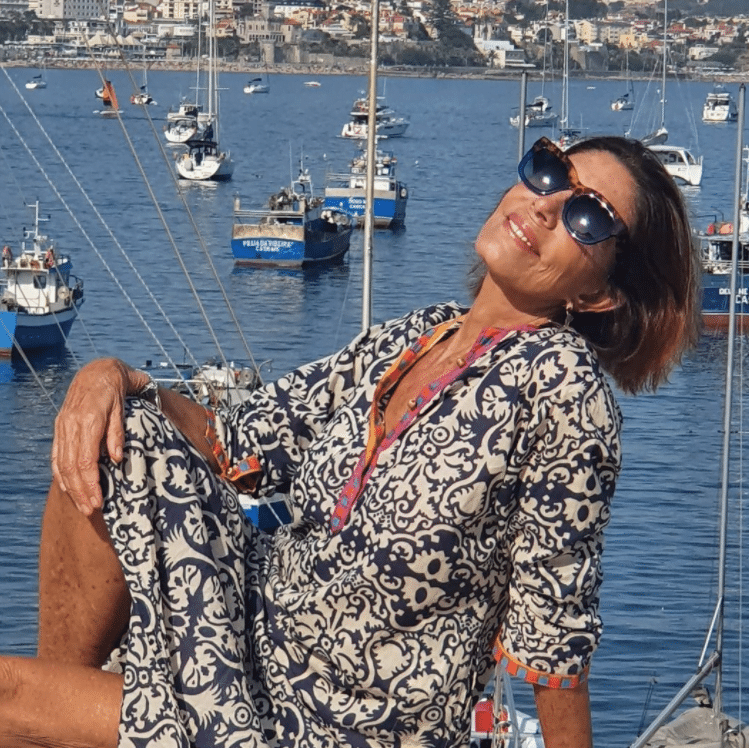 Mila Moreira de óculos escuros e vestido estampado. Atrás, cenário de barcos e do mar.