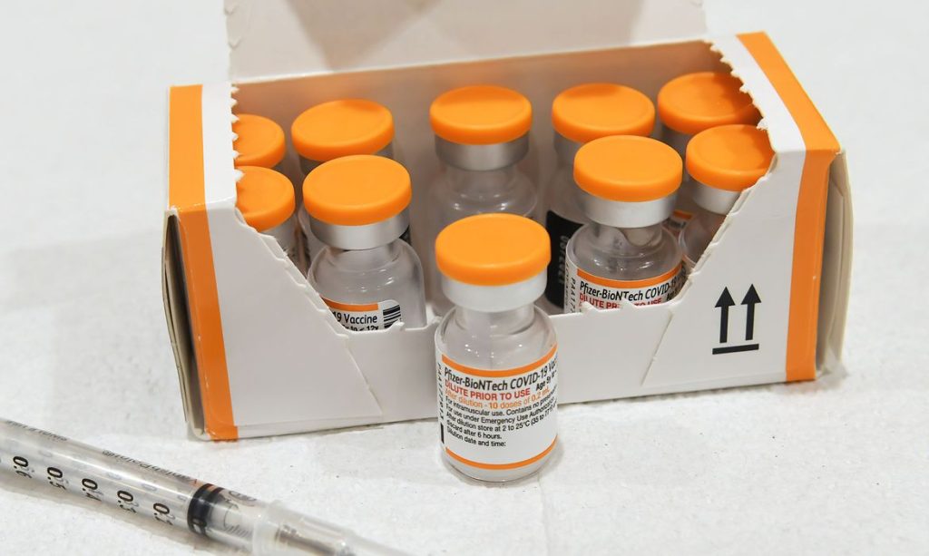 Frascos da vacina da pfizer com tampas na cor laranja. No canto esquerdo parte de um seringa.