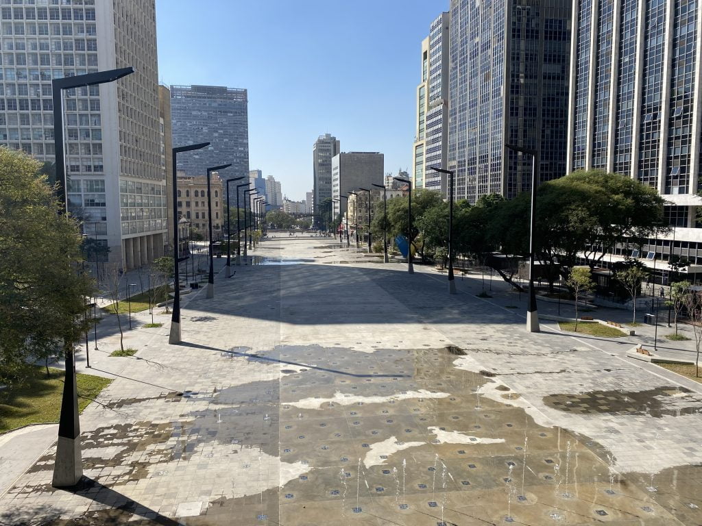 Foto panorâmica do Vale do Anhangabaú, reformado, em São Paulo. Jatos dágua que saem do chão. Luminárias em cor preta. Árvores e prédios las laterais do vale.