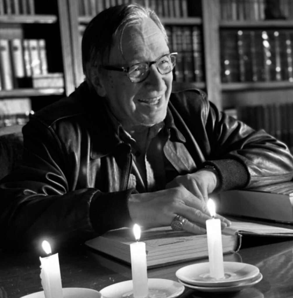 Escritor Olavo de Carvalho em foto preto e branco; aparece sentado à mesa, com os braços apoiados em um livro. Diante dele, três velas acesas. O escritor está sorrindo. Ele usa óculos de grau e jaqueta. Ao fundo, livros em uma biblioteca.