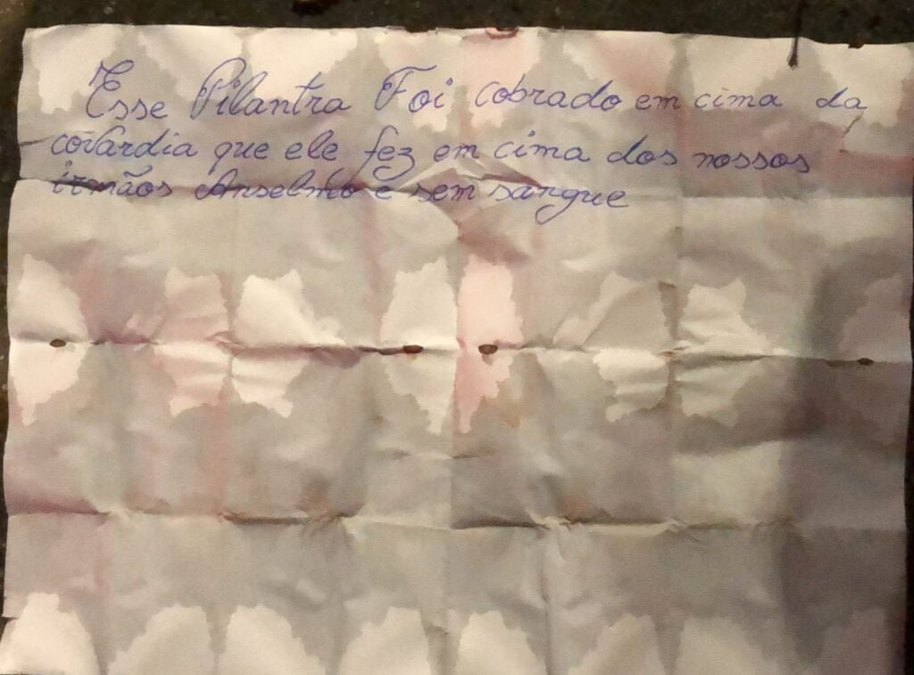 Pedaço de papel traz texto escrito com caneta azul: "Esse pilantra foi cobrado em cima da covardia que ele fez em cima dos nossos irmãos Anselmo e sem sangue"