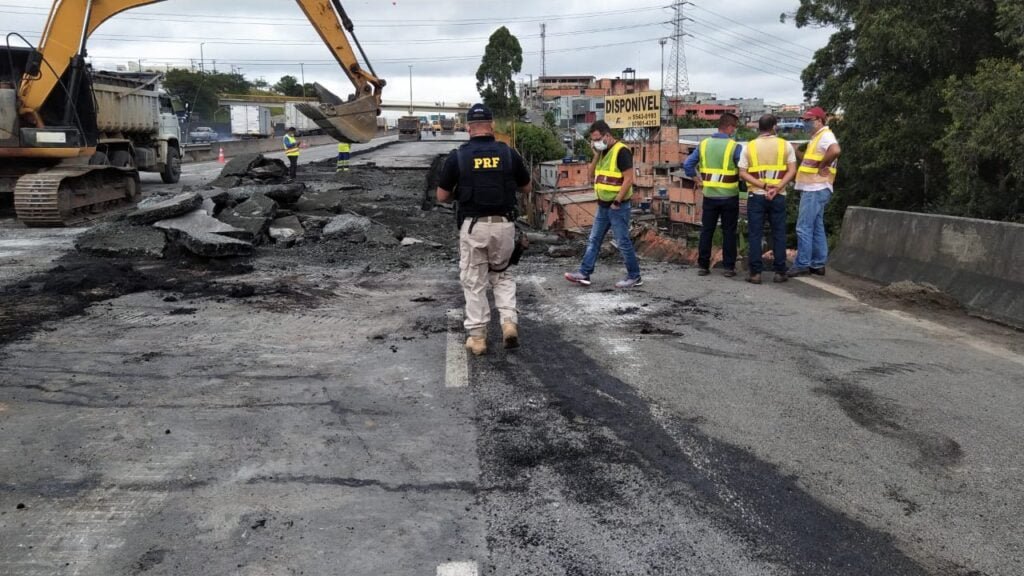 Operários e técnicos analisam cratera aberta na rodovia. Máquina retroescavadeira posicionada sobre o asfalto.