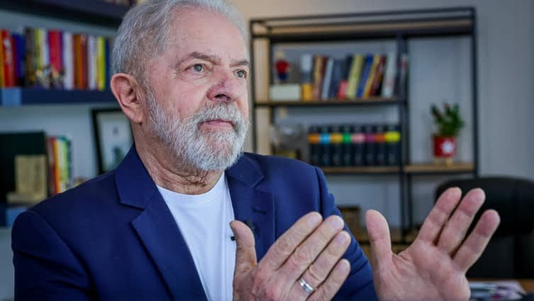 Luiz Inácio Lula da Silva, de terno e sem gravata, aparece olhando para a esquerda enquanto segura as duas mãos não altura do peito, como se apontasse um caminho.