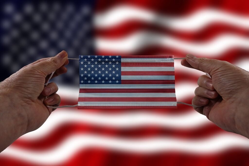 Bandeira dos Estados Unidos ao fundo, enquanto na frente duas mãos de uma pessoa branca seguram uma máscara em formato da bandeira americana.