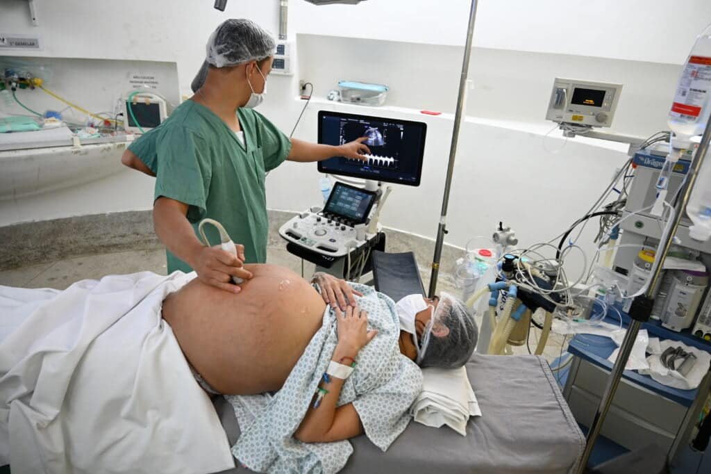 Gestante deitada, com a barriga exposta, enquanto médico observa no ultrassom os bebês em uma tela de computador. 