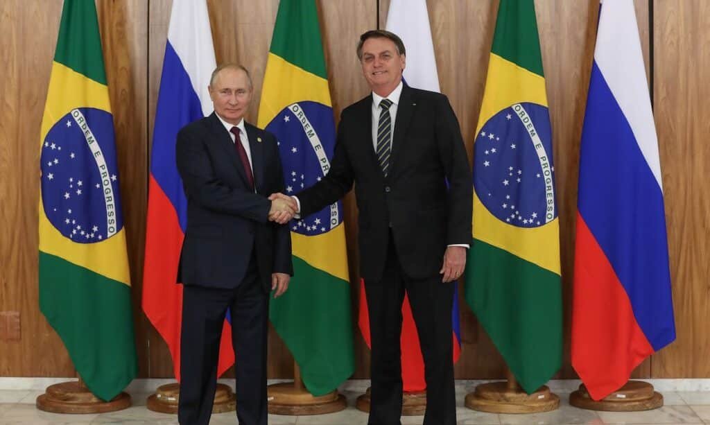 Vladimir Putine Jair Bolsonaro se cumprimentam dando a mão diante de bandeiras dos dois países.