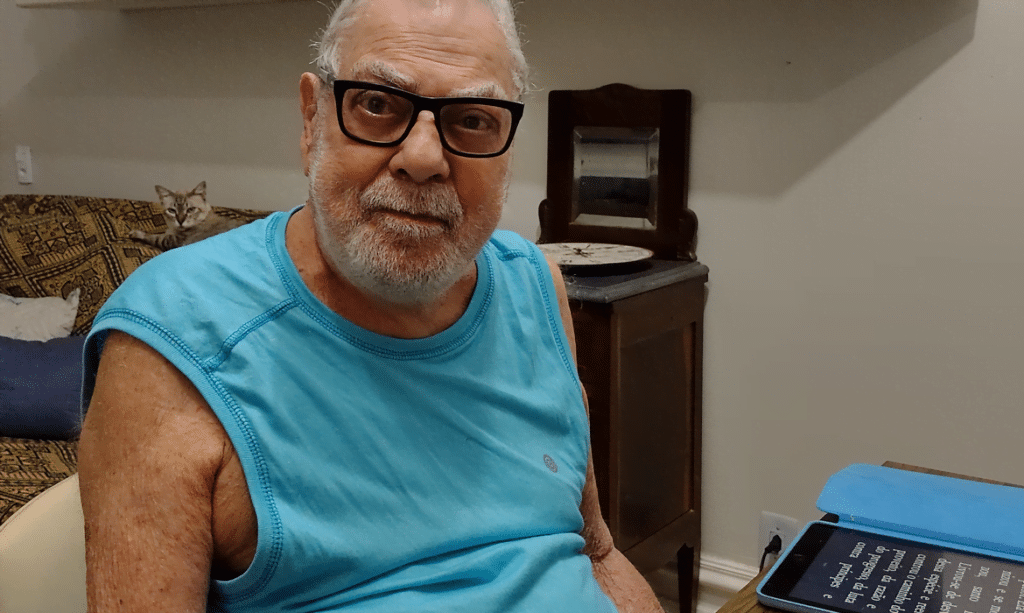 Isaac Bardavid, homem de pele clara, usa óculos com armação marcante, de linhas retas, barba e cabelos brancos, poucos cabelos na cabeça. Está sentado, de camiseta regata azul, olhando posando para foto.