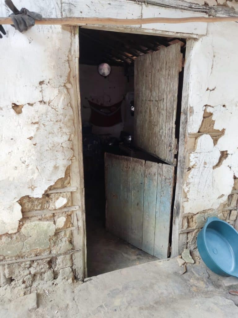 Foto mostra a porta de entrada da casa da familia. Local simples, com parede sem parte do reboco, uma bacia azul encostada na parede do lado de fora e a porta, com duas divisórias, uma na parte de cima e outra na parte baixo. A porta está aberta.
