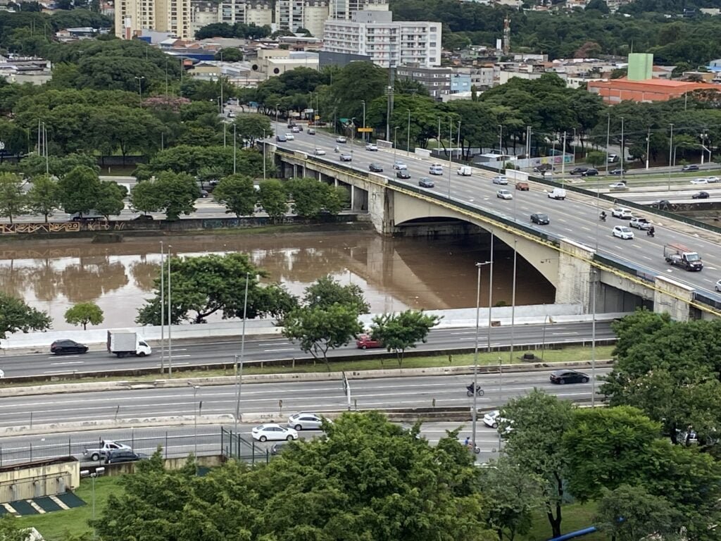 Foto aérea mostra ponte da Casa Verde sobre o rio Tietê com nível bem elevado. Casos passam pela ponte e pelas pistas da Marginal Tietê às margens do rio. 