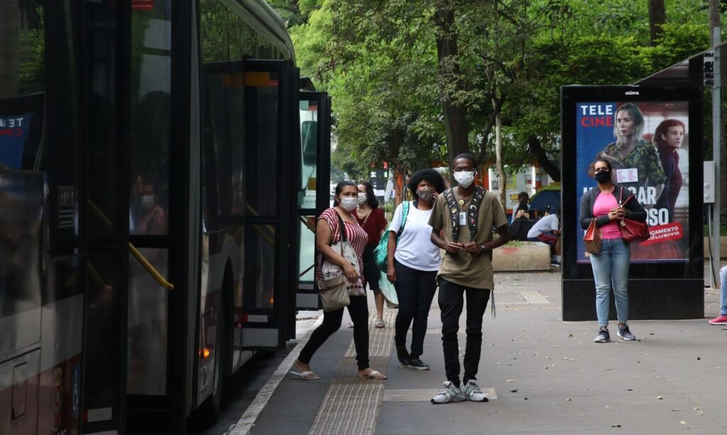 Passageiros descem de ônibus usando máscara de proteção facial enquanto outros passageiros aguardam no ponto também com máscara.