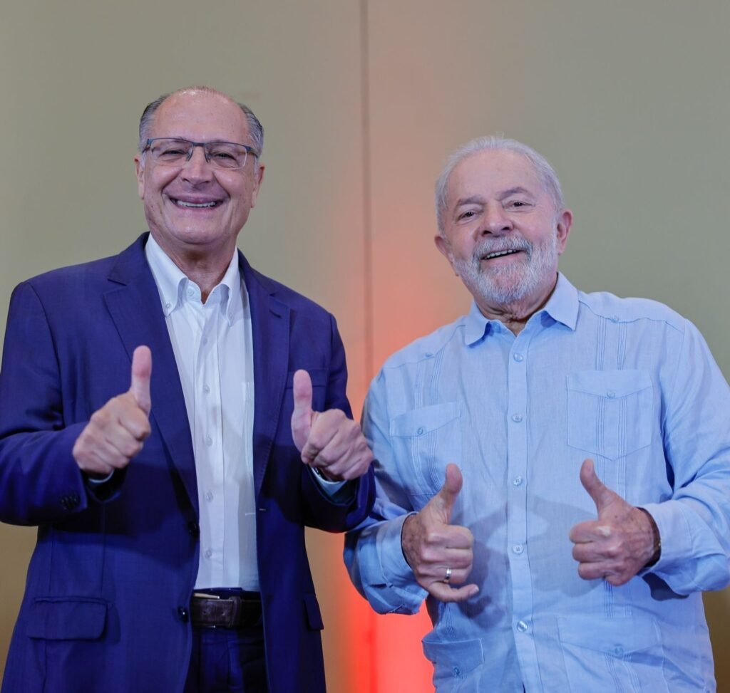 Geraldo Alckmin usando blazer e fazendo sinal de jóia com as duas mãos. Luiz Inácio Lula da Silva, ao lado de Alckmin, também faz jóia com as duas mãos. Ele usa uma camisa social azul clara. 