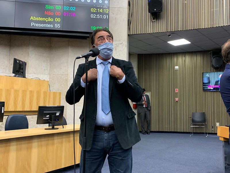 Adilson Amadeu, de terno e gravata, usando máscara, discursa ao microfone da Câmara Municipal enquanto aponta os dois dedos indicadores para si mesmo.