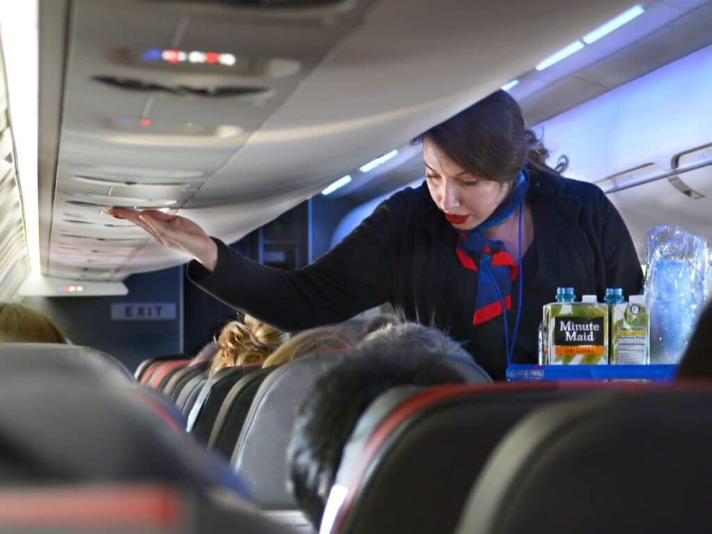 Mulher comissária de bordo conversa com passageiro enquanto empurra carrinho com bebidas.
