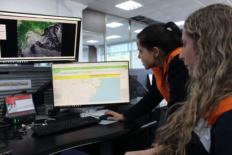 Duas mulheres meteorologistas observam tela de computador com radar meteorológicos. 