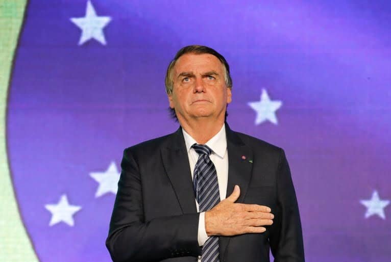 Jair Bolsonaro diante de uma bandeira do Brasil, no telão, com a mão direita sobre o peito.