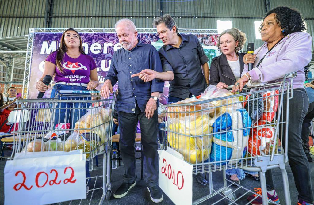 Lula e Fernando Haddad mostram dois carrinhos de compras de supermercado com alimentos dentro. Em um, mais vazio, está a placa escrita 2022. No outro, cheio, a placa escrita 2010. Duas mulheres aparecem empurrando os carrinhos.