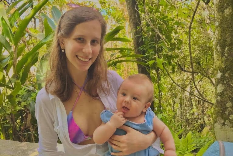 Jéssica Mayara Ballock sorri para foto com o filho no colo. Bebê aparece com a mãozinha na boca.