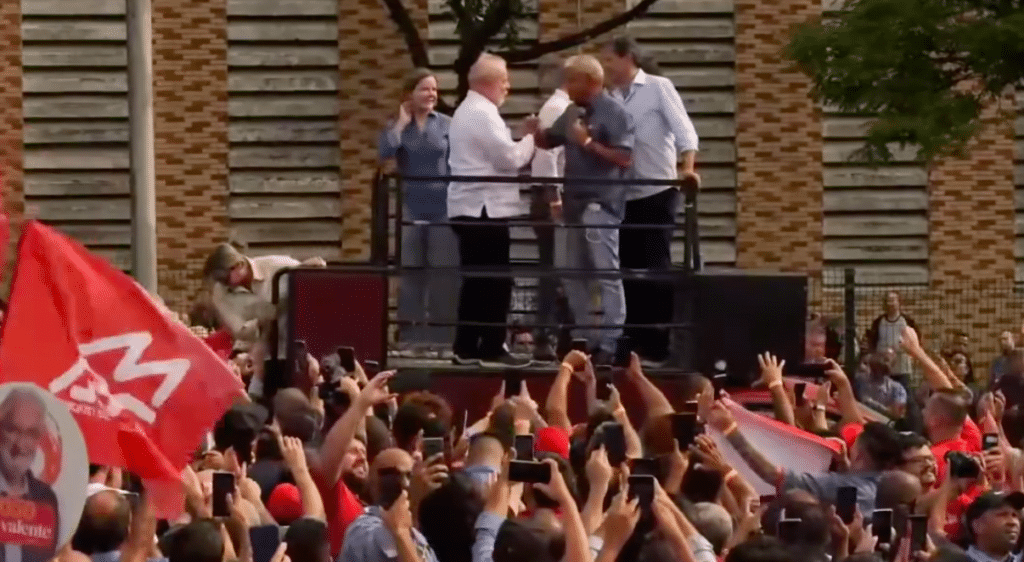 Lula em cima de palanque com outros petistas. Ele cumprimenta outro homem que está com o microfone na mão. Lula usa camisa branca. Na parte de baixo da foto aparecem mãos e celulares projetados para fazer imagens, além de uma bandeira vermelha.