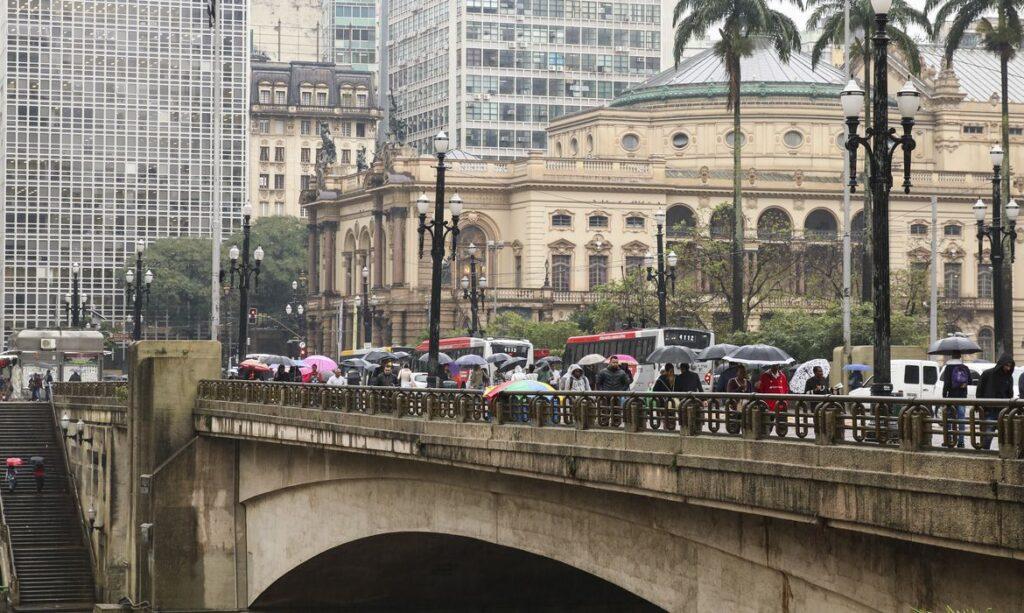 Imagem mostra pessoas caminhando sobre o viaduto do Chá, em São Paulo, com guarda-chuva.