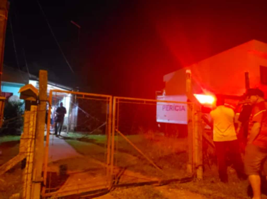 Foto mostra entrada da casa onde crime aconteceu. Placa, no alto, traz o nome "perícia". Pessoas, do lado de fora, olham na direção da casa.