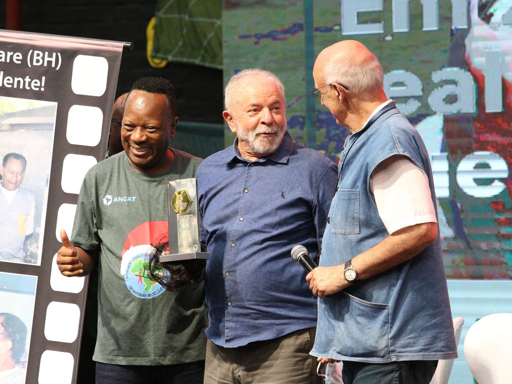 Lula aparece ao lado do Padre Júlio Lanceloti e de um dos convidados do evento sobre o palco. Lula segura uma placa com a logomarca do evento.