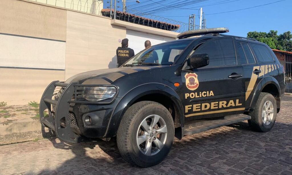 Viatura da PF estacionada em frente a uma casa. Na calçada, dois agentes aparecem de costas para a foto olhando para o portão de entrada da residencia.