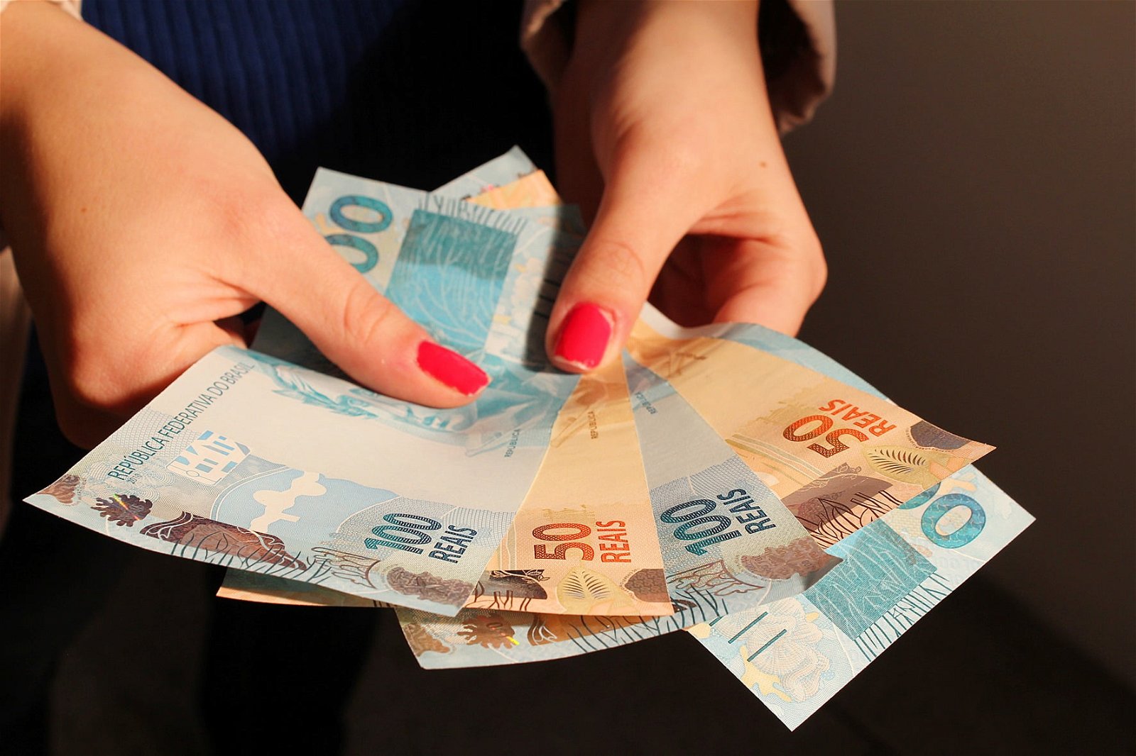Mãos de uma pessoa branca, com unhas pintadas de vermelho, seguram cédulas de dinheiro.