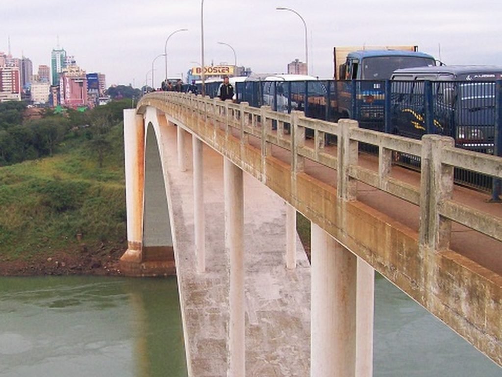 Ponte da amizade com veículos parados em cima da estrutura. No canto esquerdo é possível ver a água do rio. Ao fundo, grama e parte da cidade no Paraguai.