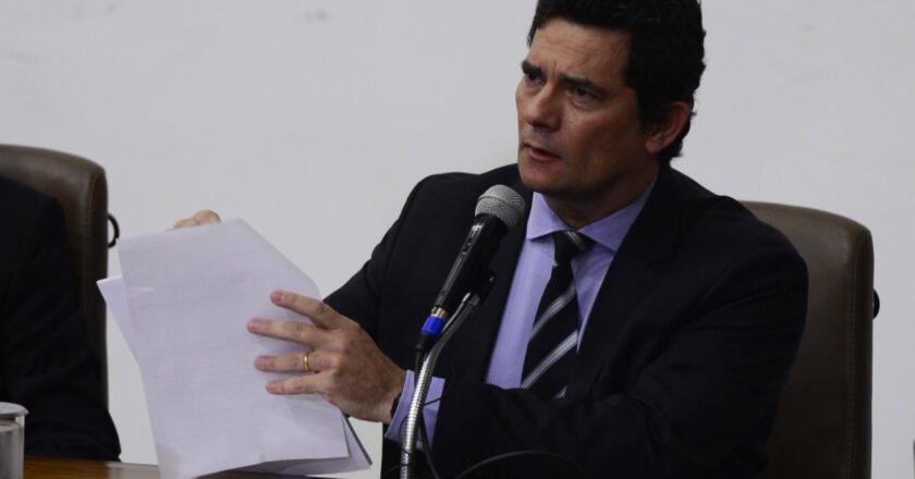 Sergio Moro segura papei enquanto fala sentado à mesa diante de um microfone.