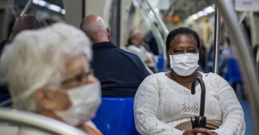 Passageiras sentadas dentro do vagão do metrô usando máscara. No primeiro plano, à esquerda, uma senhora com cabelos brancos e óculos de grau aparece de lado. Ao fundo, de frente para a câmera, uma senhora segura o guarda chuva enquanto também usa máscara facial.