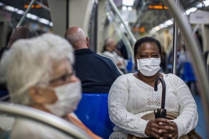 Passageiras sentadas dentro do vagão do metrô usando máscara. No primeiro plano, à esquerda, uma senhora com cabelos brancos e óculos de grau aparece de lado. Ao fundo, de frente para a câmera, uma senhora segura o guarda chuva enquanto também usa máscara facial. 