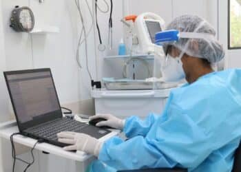 Cientista em laboratório da Fiocruz, usando equipamentos de proteção individual no corpo e na cabeça, além de luvas, utiliza computador dentro do laboratório.