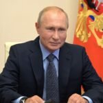 Vladimir Putin, homem de pele clara, poucos cabelos loiros, veste terno e gravata. Atrás dele aparece parte de uma bandeira.