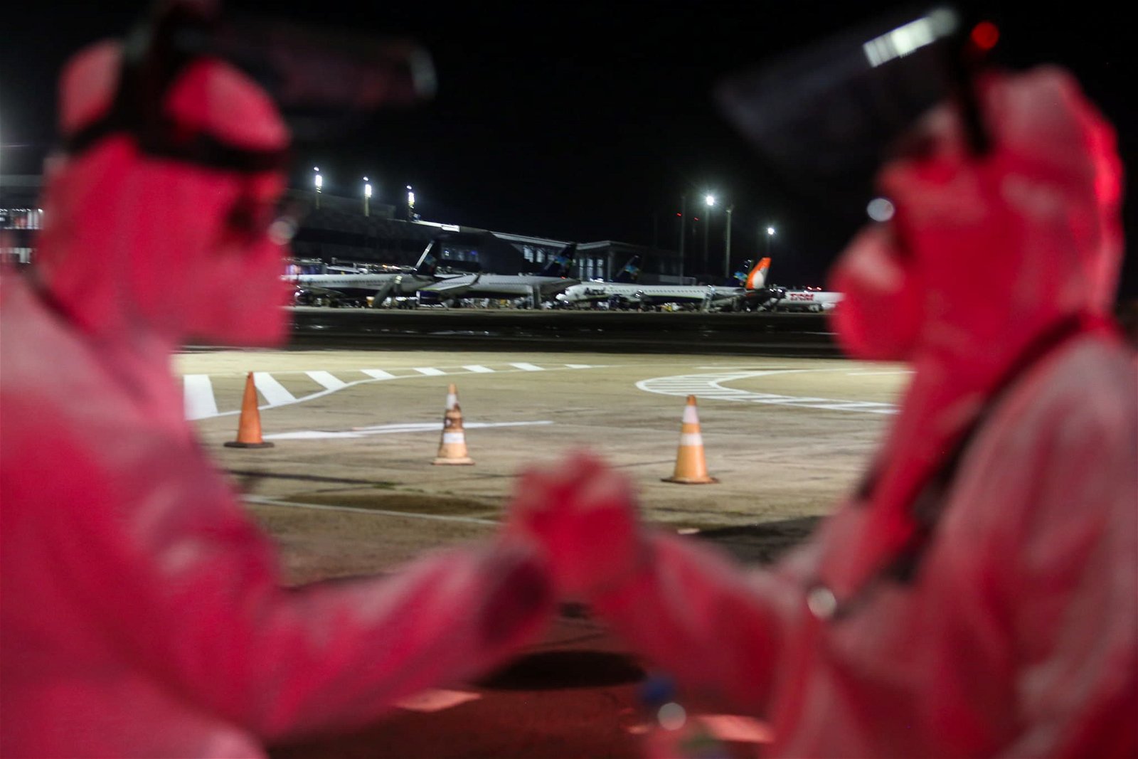 Duas pessoas, vestindo roupa especial anti-infecção se cumprimentam. Ao fundo, aviões estacionados no aeroporto e cones de sinalização na pista do aeroporto.