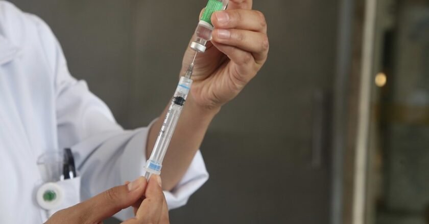 Agente de saúde usa seringa e agulha para extrair a vacina de dentro do frasco. Imagem mostra as mãos do agente e parte do jaleco branco. Ao fundo uma parede desfocada.