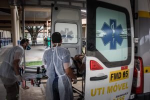 Paciente é colocado em ambulância por dois socorristas que usam equipamentos de proteção pessoal