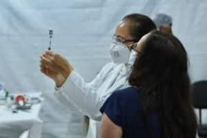 Enfermeira prepara vacina que será aplicada em mulher, que acompanha o procedimento usando máscara.