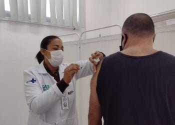 Agente de saúde, de máscara e jaleco branco, aplica vacina no braço de homem.