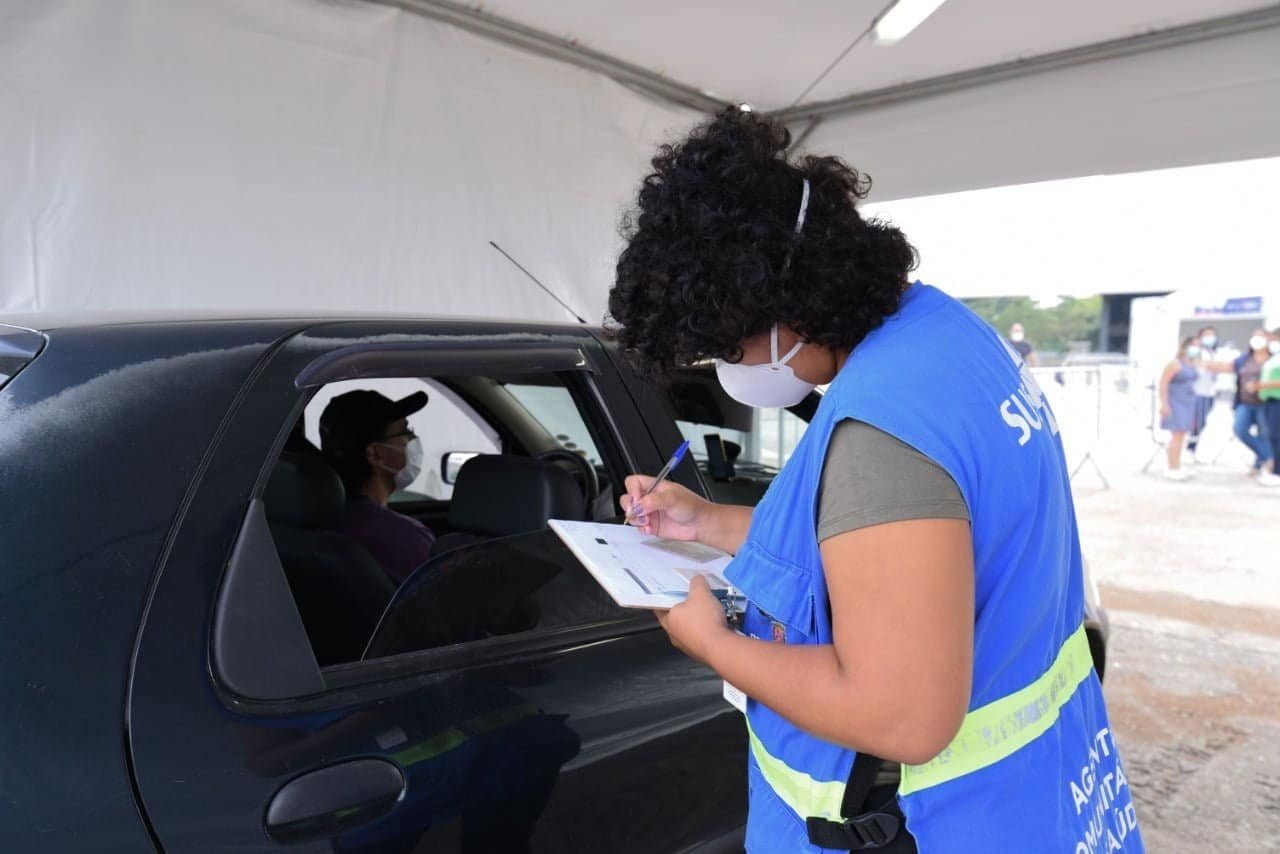 Agente de saúde anota dados em uma folha na prancheta enquanto conversa com paciente que está dentro do carro esperando pela vacina