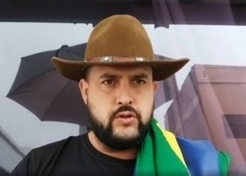 Zé Trovão, homem branco, com bigode e barba, usa chapéu marrom, a bandeira do Brasil sobre o ombro esquerdo e veste camiseta preta.