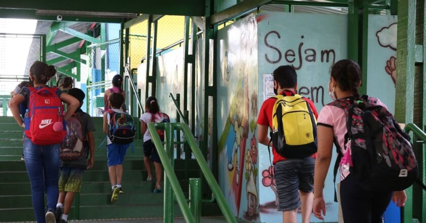 Alunos com mochilas nas costas sobem escada pintada de verde, com corrimão verde, na entrada de uma escola. Na parede, partes de palavras escritas em várias cores.