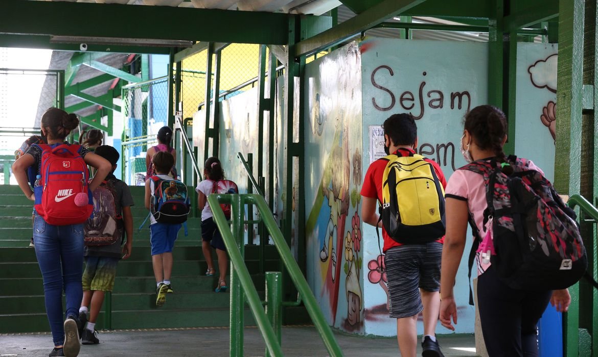 Alunos com mochilas nas costas sobem escada pintada de verde, com corrimão verde, na entrada de uma escola. Na parede, partes de palavras escritas em várias cores.