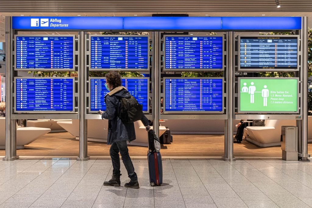 Passageiro caminha puxando sua mala no aeroporto, na Alemanha, usando máscara. Ao fundo, paineis indicam os voos.