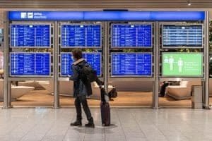 Passageiro caminha puxando sua mala no aeroporto, na Alemanha, usando máscara. Ao fundo, paineis indicam os voos.