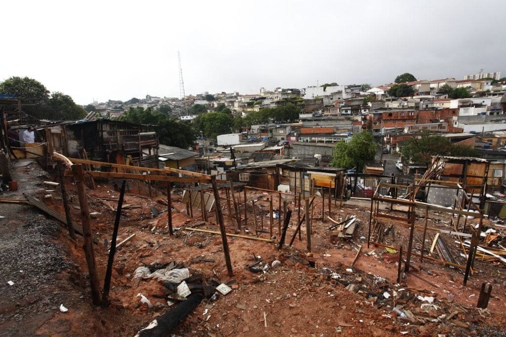 Área atingida pelo fogo. Pedaços de madeiras usados para montar barracos ainda fincadas no solo. Ao fundo a cidade de São Paulo, com casas de alvenaria.