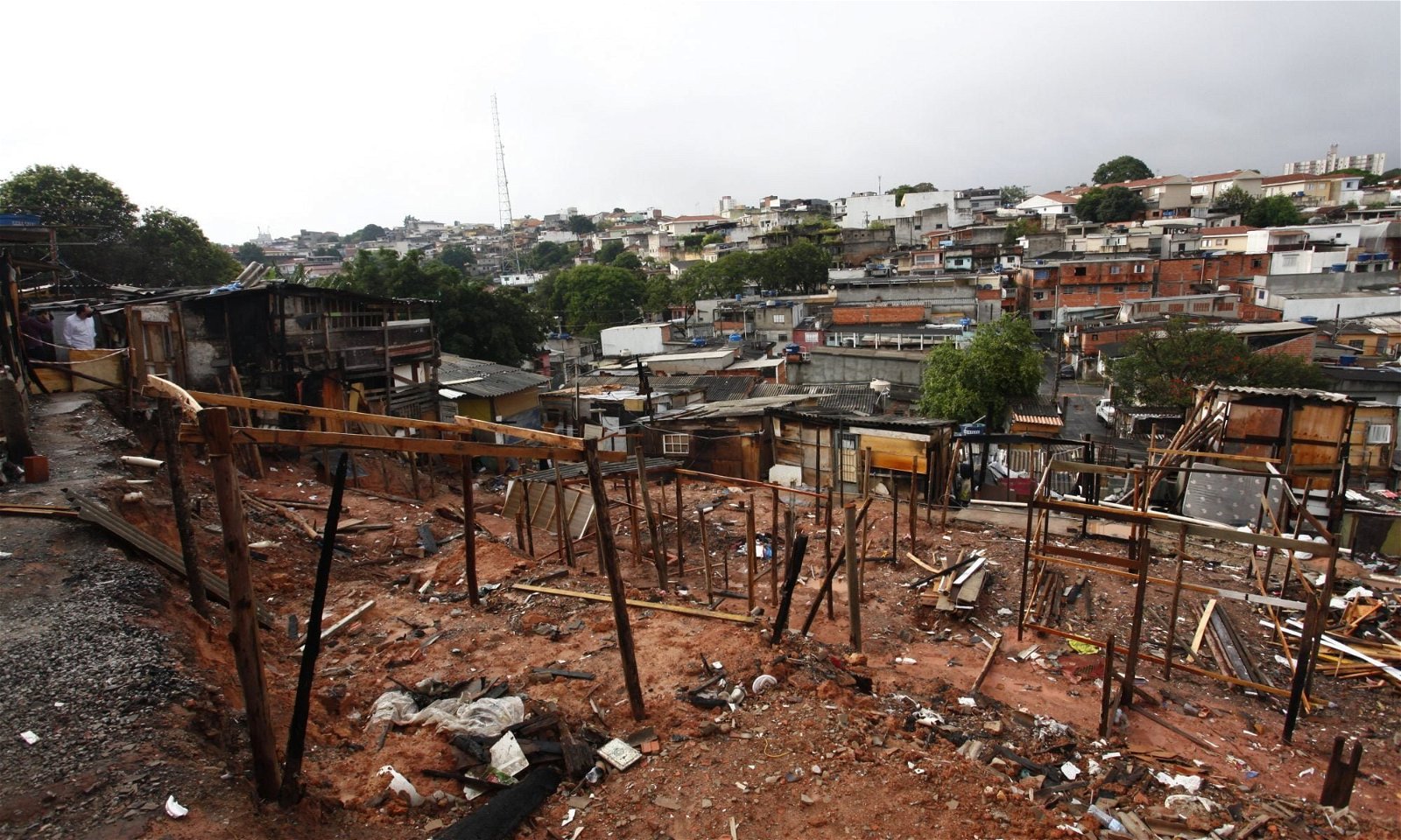 Área atingida pelo fogo. Pedaços de madeiras usados para montar barracos ainda fincadas no solo. Ao fundo a cidade de São Paulo, com casas de alvenaria.